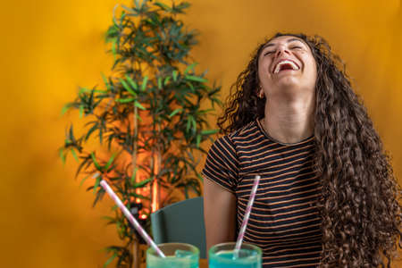 beautiful woman with long curly hair laughing in front of two blue drinks on colored backgroundの写真素材
