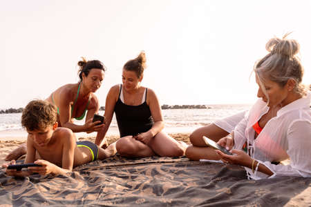 family on vacation using smartphone sitting relaxed on the beach at sunset - summer vacation conceptの写真素材