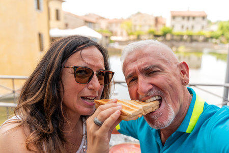 beautiful middle aged couple taking a funny selfie while eating a sandwich in a historic italian village - travel and vacation conceptの写真素材