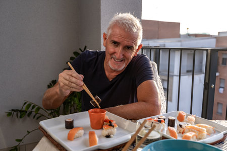 attractive middle-aged man eating while sitting at a table laid with Chinese take away food in a city balconyの写真素材