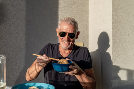 attractive middle-aged man have fun while eating sitting at a table laid Chinese take away food in...