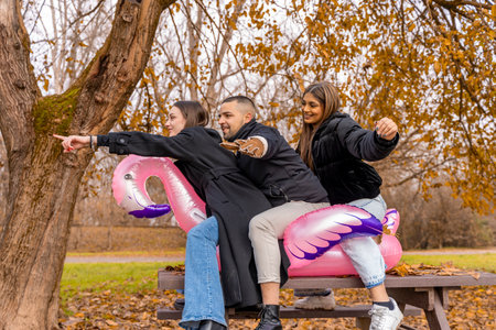 small group of young friends having fun riding an inflatable pink flamingo over a wooden table in a public parkの写真素材