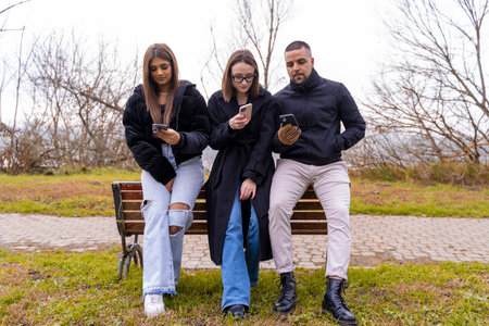 small group of young friends having fun using smart phone leaning on a bench in a public park in...