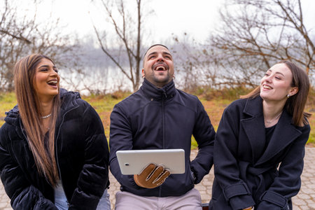 three young male and female friends using tablet having fun sitting on an iron bench in a public...