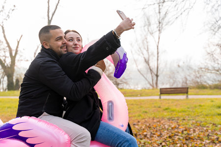 portrait of two young lovers taking a selfie having fun riding an inflatable pink flamingo in a public park in winterの写真素材