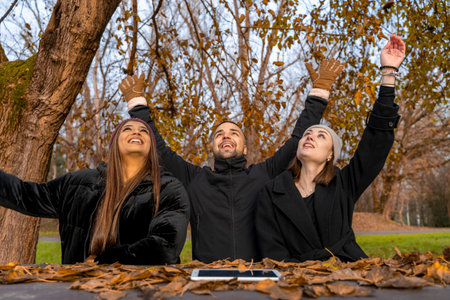 three young friends male and female in winter clothes raising hands up sitting at a table in a public park in winterの写真素材