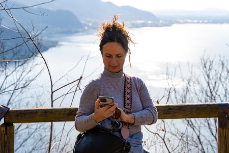 pretty smiling middle aged woman using mobile phone leaning on wooden fence on lakeside pathの写真素材