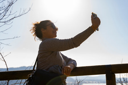 pretty smiling middle aged woman taking a selfie leaning on a wooden fence on a lakeside path