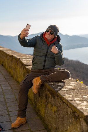 handsome middle-aged man in winter clothes and sunglasses taking a selfie sitting on a stone wall in front of a beautiful lake viewの写真素材