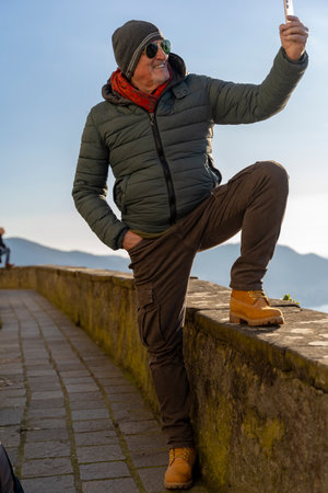 handsome middle-aged man in winter clothes and sunglasses taking a selfie sitting on a stone wall in front of a beautiful lake viewの写真素材