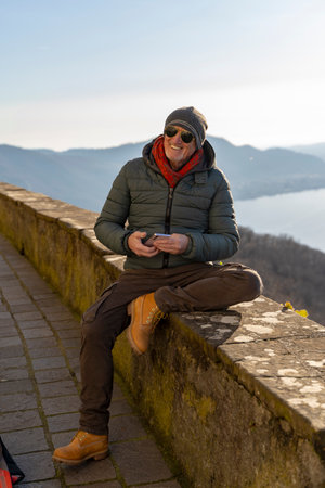 handsome middle aged man in winter clothes using mobile phone sitting on a stone wall in front of a beautiful lake viewの写真素材