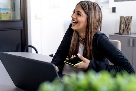 young smiling business woman working typing on laptop keyboard and talking on mobile phone in modern officeの写真素材