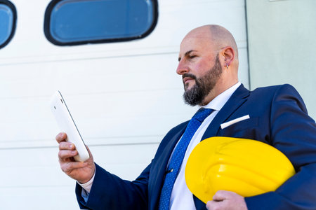 bearded businessman working using mobile phone outdoors wearing protective yellow helmet and transparent work glassesの写真素材