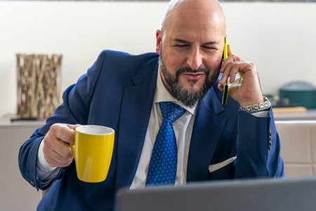 portrait of smiling businessman talking on mobile phone in front of laptop during coffee break in an officeの写真素材