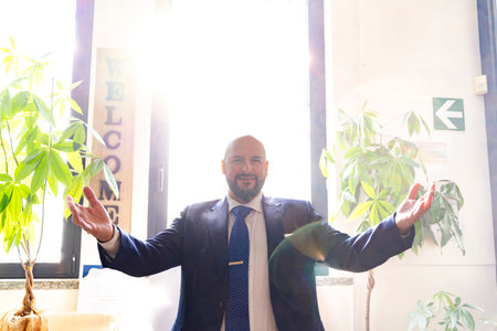 benevolent smiling businessman in elegant suit opening his arms surrounded by divine light in his officeの写真素材