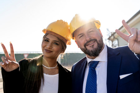 portrait of two smiling young businessmen, man and woman, showing victory sign outdoors wearing yellow safety helmetの写真素材