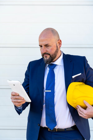 portrait of bearded businessman working outdoors using tablet wearing yellow protective helmetの写真素材