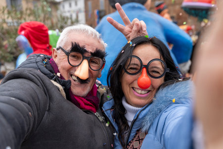 middle aged couple taking a selfie having fun with masks and confetti during a carnival parade in the streetの写真素材