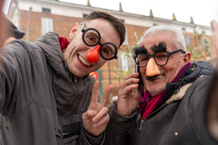 couple of male friends taking a selfie wearing funny masks under a rain of confetti during a carnival parade in the streetの写真素材