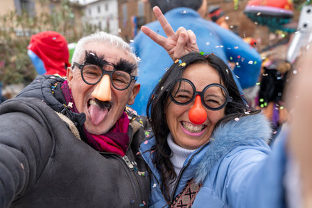 middle aged couple taking a selfie having fun with masks and confetti during a carnival parade in the streetの写真素材