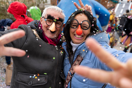 middle aged couple taking a selfie having fun with masks and confetti during a carnival parade in the streetの写真素材