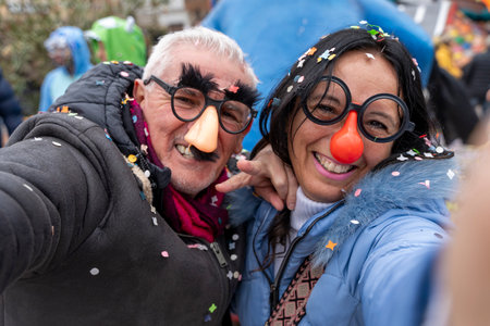 middle aged couple taking a selfie having fun with masks and confetti during a carnival parade in the streetの写真素材