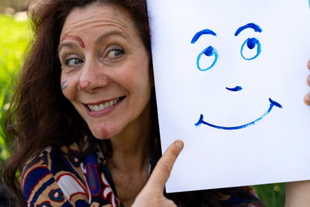 beautiful smiling painter woman with red hair making funny faces showing a stylized smiley portrait on a white sheetの写真素材