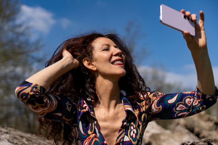 smiling red-haired woman taking a funny selfie sitting on a big log outdoorsの写真素材