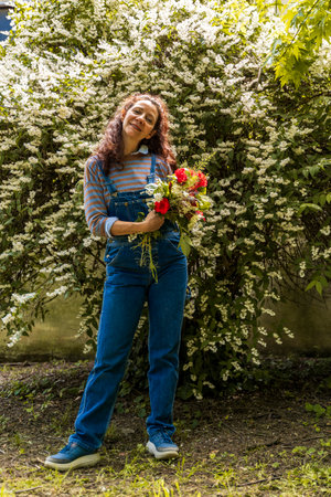cute smiling woman with red hair showing a bunch of colorful flowers standing in front of a tree with white flowersの写真素材