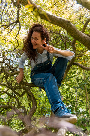 beautiful smiling woman with red hair showing peace symbol with her hand on twisted branches of a holm oak treeの写真素材