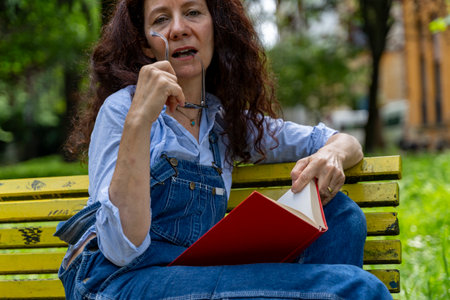 middle aged woman sitting on a yellow bench looking into the camera reading a book with a red coverの写真素材