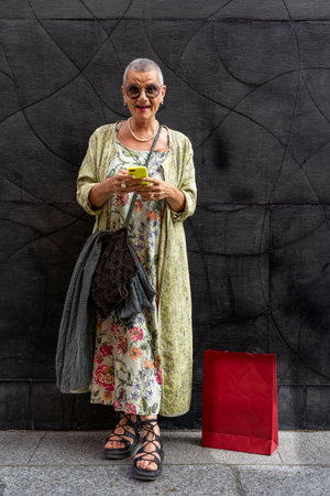 elegant smiling mature woman with shaved gray hair texting on cell phone in front of black wall