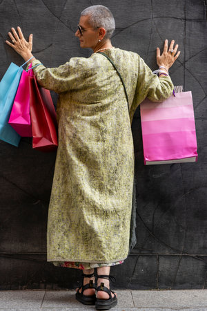 elegant mature woman with colorful shopping bags posing with her back turned in front of a black wallの写真素材