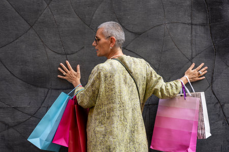 elegant mature woman with colorful shopping bags posing with her back turned in front of a black wallの写真素材