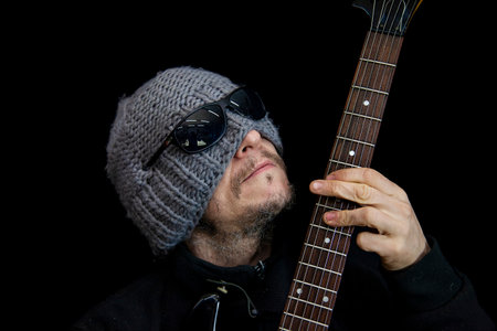 Man with beanie and sunglasses covering eyes, intensely playing electric guitar on a dark background