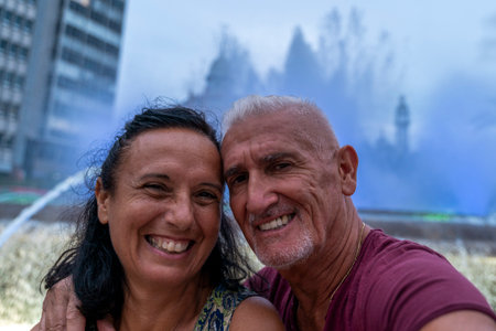 beautiful smiling middle-aged couple on vacation taking a selfie in front of a fountain with colored...