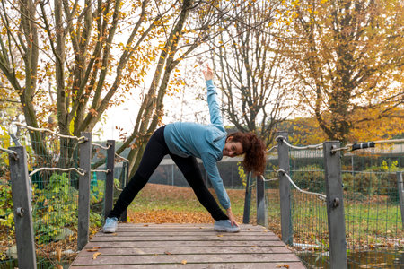 Woman performing a yoga pose on a wooden bridge in an autumn park for wellness