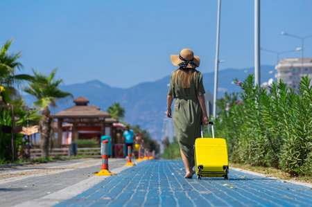 A woman in a green dress walks with a yellow suitcase along the sidewalk.の写真素材