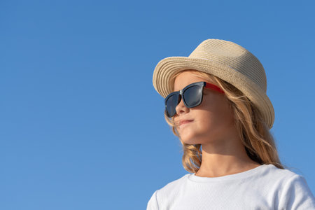 Close-up portrait of a young girl in sunglasses and a hat against the blue sky. View from a low angle.の写真素材