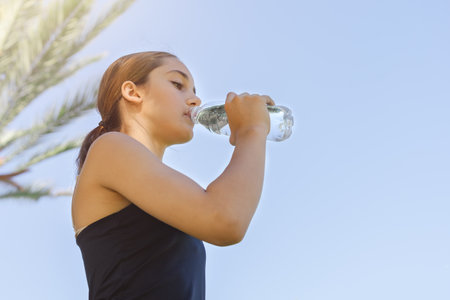 Low angle view of a young girl drinking water from a plastic bottle against a blue sky.の写真素材