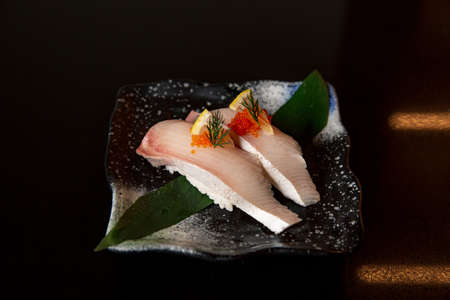 Kingfish sushi with tobiko flying fish roe in the fine dining Japanese restaurant, sashimi, nigiri and raw fish with a leaf plating in black scene reflection table backgroundの写真素材