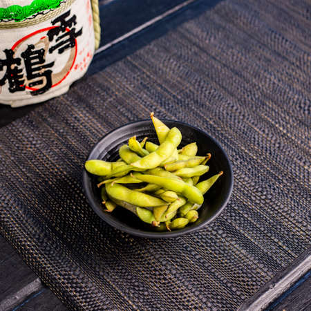 Edamame soybeans in the traditional Japanese ramen restaurant, with a black plate on a black mat and Japanese fonts sake barrel translation: Snow Craneの写真素材