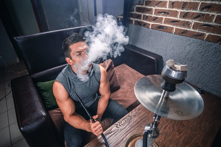 Young handsome man blowing clouds of smoke in the shisha hose.の写真素材