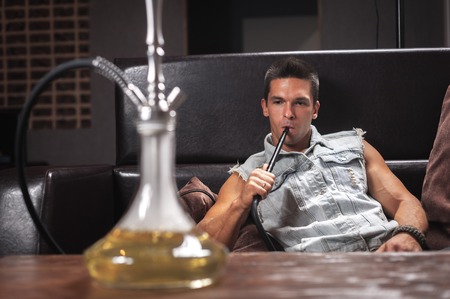 Young handsome man blowing clouds of smoke in hookah cafe.