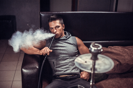 Sexy young man smoking a hookah and smoke issues from the mouth at arabic restaurant.