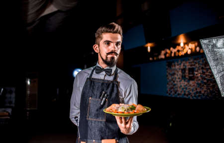 Professional friendly waiter carries appetizing dish on a black background.