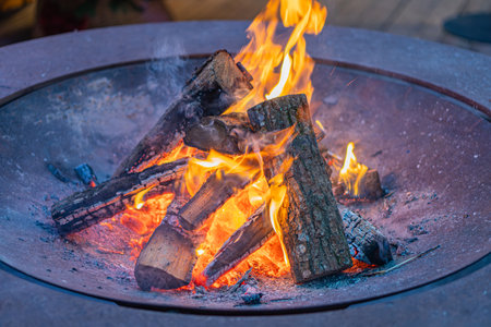 Image depicts a nighttime fire pit, surrounded by burning logs within a circular metal frame The...