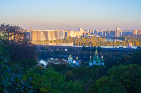 Panoramic cityscape view, possibly sunrise or sunset, lush green trees, dome-like buildings, body of...