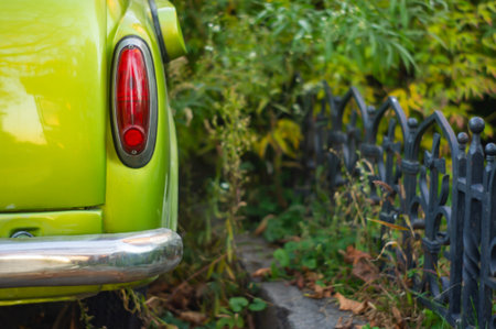 An old, yellowgreen vintage car distinctive rounded shape, no distinguishing marks and a weathered metal fence intricate design, small leaves growing through gaps in a candid photographの写真素材