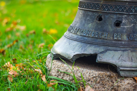 A weathered metal bell, mounted on a concrete base outdoors, with inscriptions and a dark patina. Surrounded by grass and fallen leaves, the background shows green foliage.の写真素材